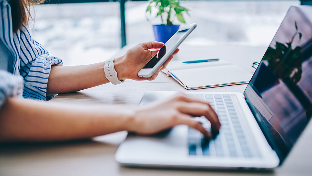 En person använder en laptop och håller en smartphone i ena handen. På skrivbordet finns en anteckningsbok, en penna och en liten växt, med en ljus och modern bakgrund.