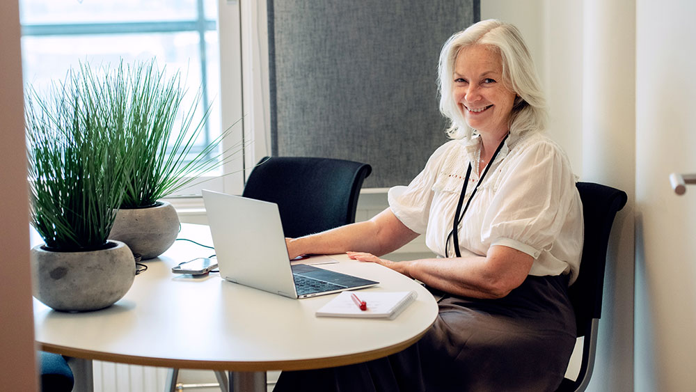 En kvinna med grått hår sitter vid ett runt bord och ler mot kameran medan hon arbetar på en laptop. På bordet ligger ett anteckningsblock, en penna och två krukväxter, vilket skapar en bekväm och välkomnande atmosfär.