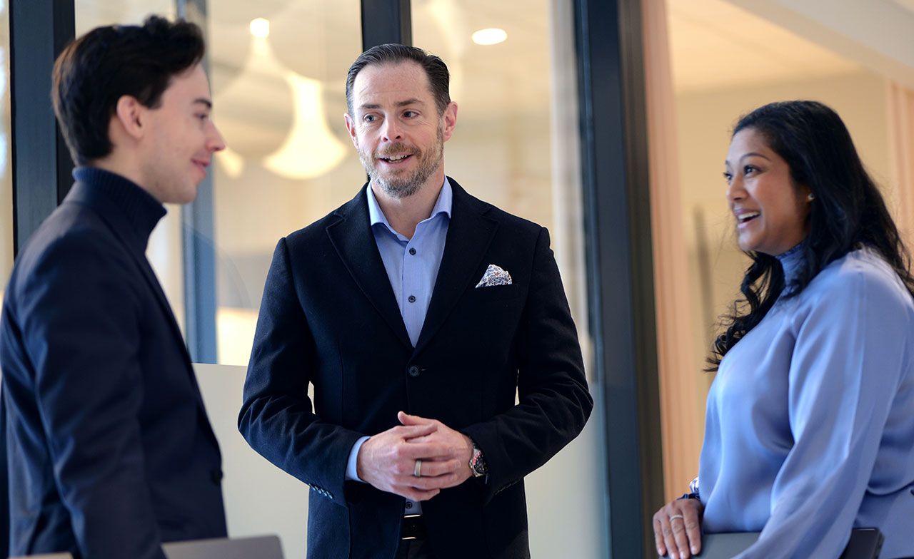 business_professionals_conversation_office_networking Tre personer i affärskläder står och samtalar i en modern kontorsmiljö. De ler och verkar ha en avslappnad och professionell dialog.