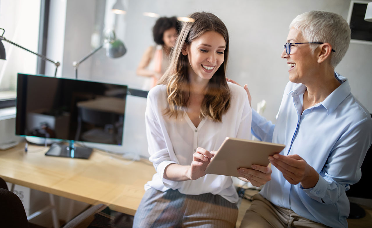colleagues_collaboration_315286633 Två kvinnor i olika åldrar samarbetar leende framför en surfplatta på ett modernt kontor. En sitter på skrivbordet, den andra står bredvid, och i bakgrunden syns ytterligare en kollega. Stämningen är positiv och stödjande.