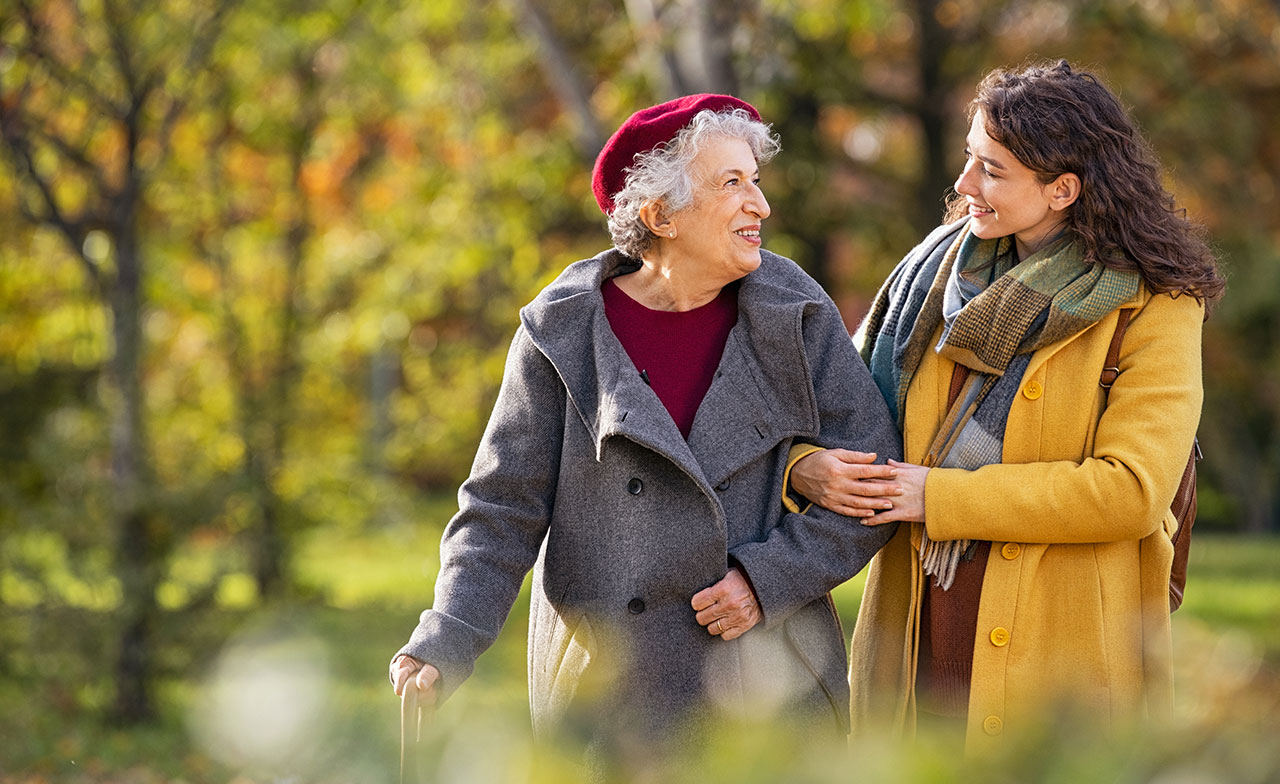 elderly_woman_walking_with_younger_caregiver_autumn_park En äldre kvinna och en yngre kvinna promenerar tillsammans i en solig park med höstfärger. De håller varandra i armkrok, ler och ser glada ut i varandras sällskap. Den äldre kvinnan bär en grå kappa och röd basker, den yngre en gul kappa och halsduk.