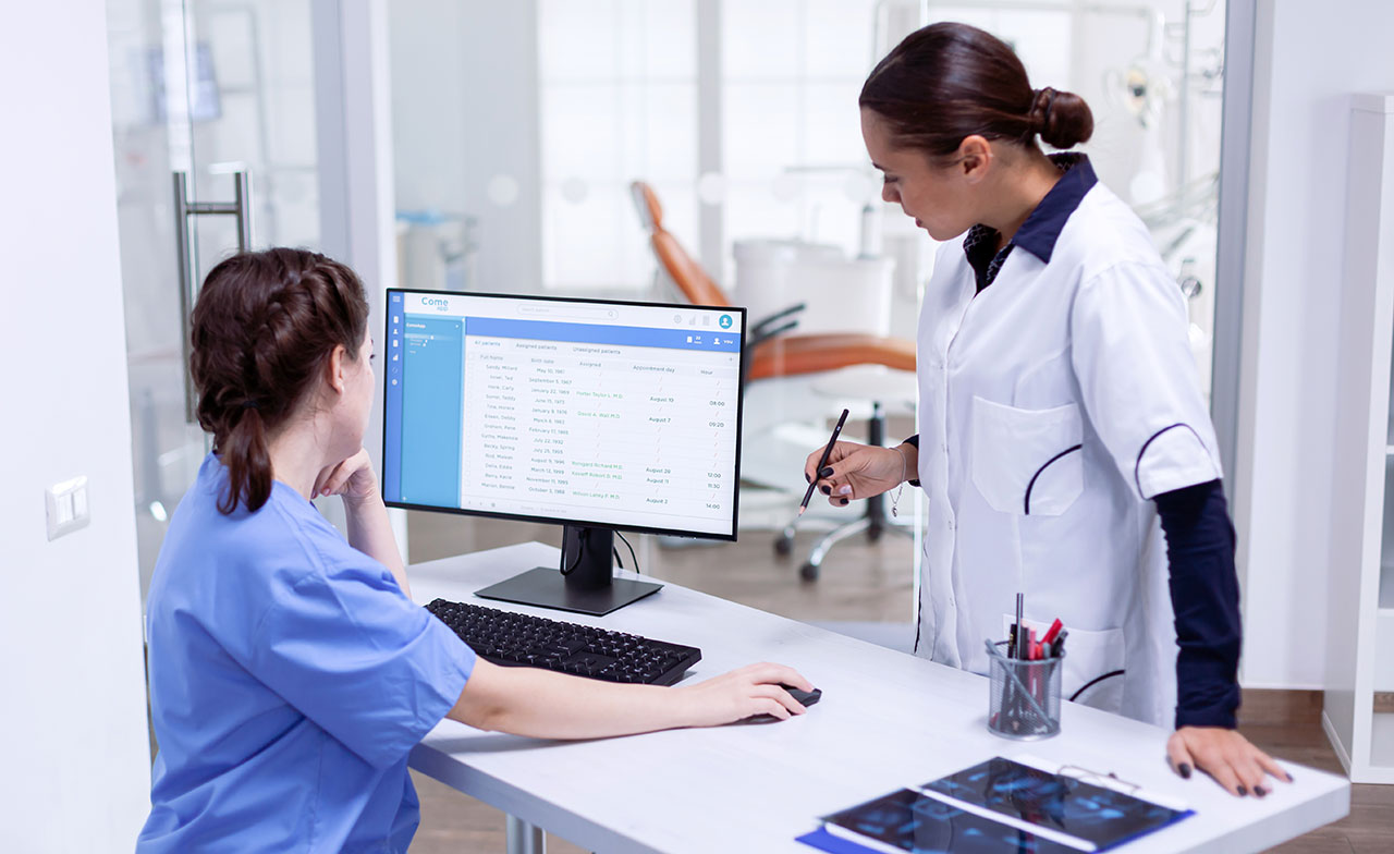 healthcare_staff_reviewing_patient_data_on_computer_392194268 Två vårdpersonal i arbetskläder står vid ett skrivbord på en klinik och granskar patientinformation på en datorskärm. En sitter vid datorn och den andra pekar på skärmen med en penna. På bordet ligger även röntgenbilder.