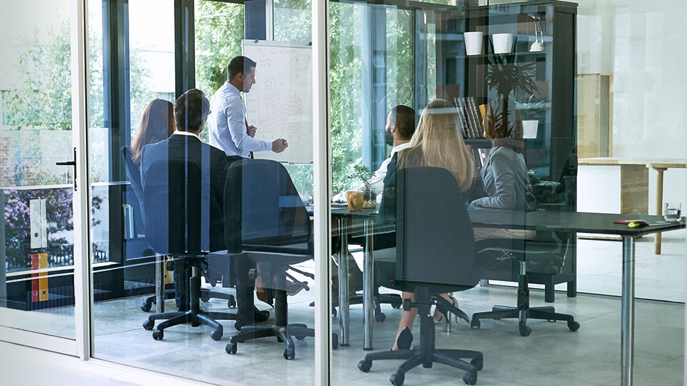 Business Consulting representanter ses i ett mötesrum genom glasväggar där en person presenterar en analys av inköpsorganisationen för sina kollegor