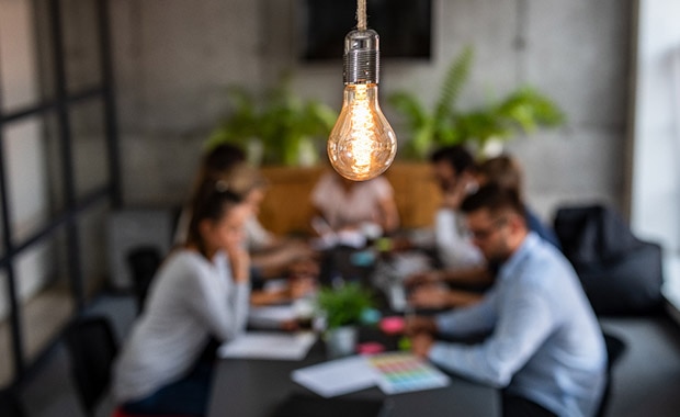 Gruppe junger Leute sitzen gemeinsam an einem Tisch, darüber hängt eine leuchtende Glühbirne Gruppe junger Leute sitzen gemeinsam an einem Tisch, darüber hängt eine leuchtende Glühbirne