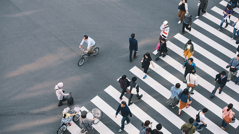 Flygbild av ett övergångsställe fullt med fotgängare som korsar gatan i olika riktningar, medan en cyklist passerar på sidan.