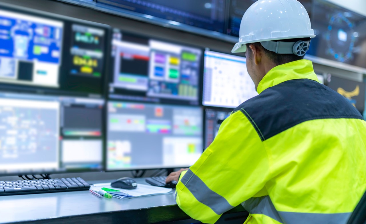 Person wearing a yellow jacket and white hardhat monitoring computer data