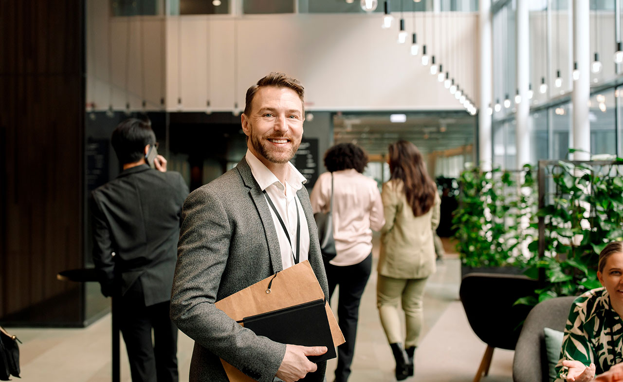 En leende man i kostym står i en modern kontorslobby med ett dokumentfodral i famnen. I bakgrunden syns andra personer i rörelse samt växter och stora fönster som släpper in dagsljus. businessman_smiling_in_modern_office_lobby