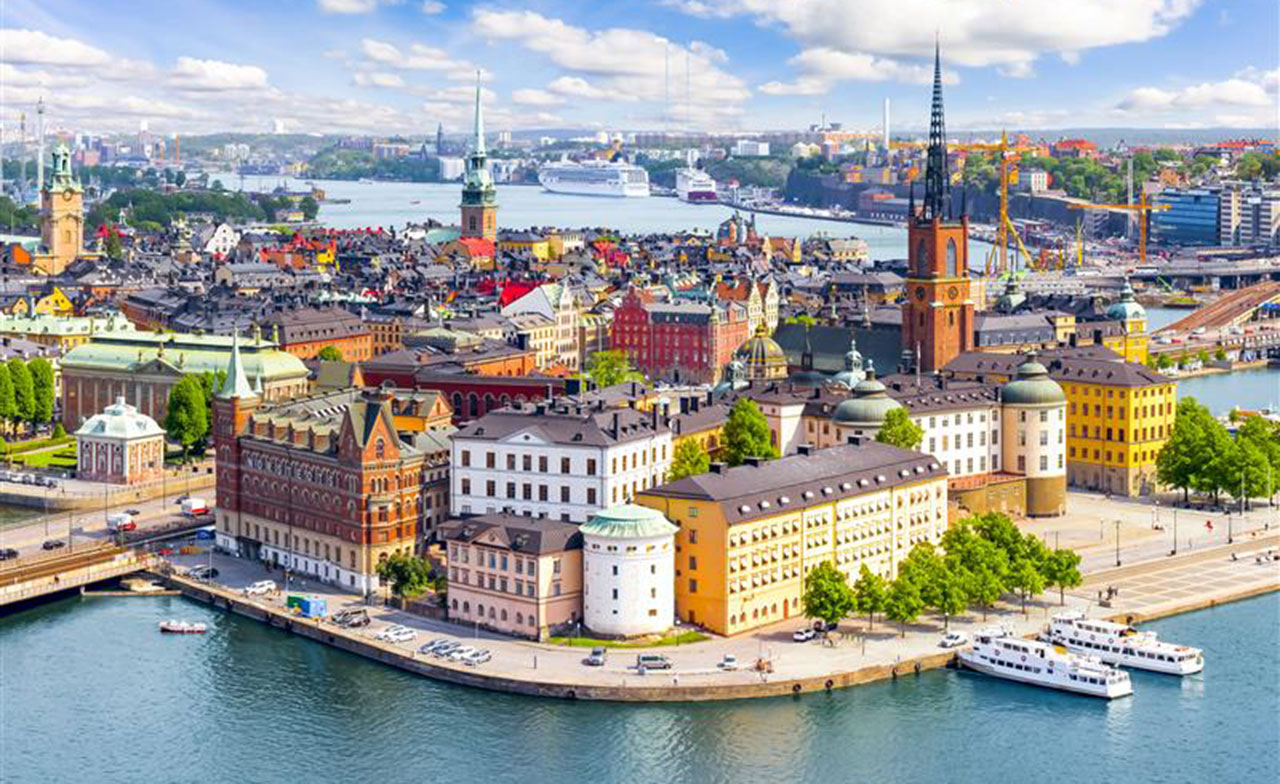  färgstark och pittoresk vy över Stockholm, Sveriges huvudstad. I förgrunden syns Riddarholmen, en historisk del av staden med sina karakteristiska byggnader och Riddarholmskyrkan med sitt spetsiga torn. Bakom syns Gamla stan med sina färgglada hus och smala gränder. I bakgrunden breder Stockholm ut sig med moderna byggnader, båtar och vatten, vilket ger en levande känsla av stadens skärgårdslandskap.