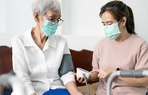Kvinnlig vårdpersonal med munskydd sitter bredvid äldre kvinnlig patient som också har munskydd och kontrollerar blodtryck