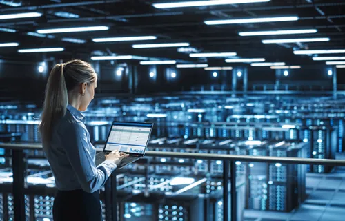 A female professional looking at a laptop while overlooking rows of servers 