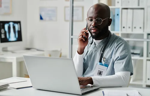 Young confident physician in medical scrubs and eyeglasses communicating with patients online and on mobile phone