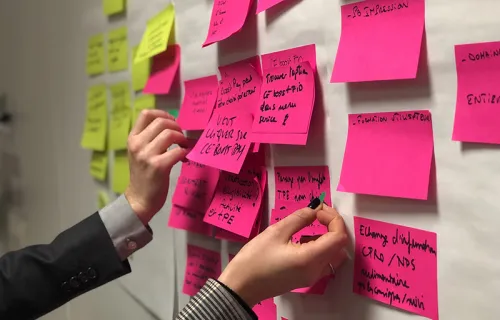 Rosa och gula post-it lappar på en white board där två personer också sträcker fram sina händer…