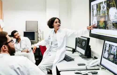 Kvinnlig läkare i vit sjukhusrock står lutad mot ett bord och vänder sig mot skärmarna bakom och pekar på patientinformation som hennes kollegor i rummet också tittar på