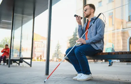 Junger blinder Mann mit Smartphone sitzt auf einer Bank und spricht.