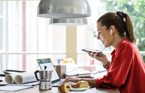 Kvinna i röd topp sitter vid sitt frukostbord i köket och jobbar med laptopen uppfälld och en…
