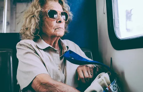 En äldre dam med solglasögon sitter på ett tåg och tittar ut genom fönstret