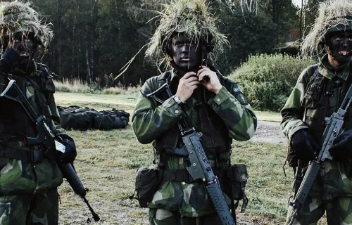 Tre militärklädda män med hjälmar och vapen samt målade i ansiktet står och tittar mot kameran ute på fältet