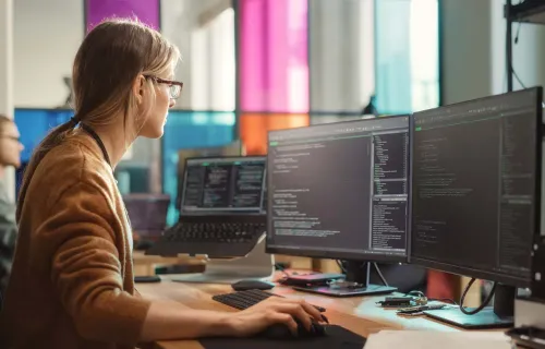 Person arbeitet an mehreren Monitoren mit Programmcode in einem Entwicklungsumfeld.