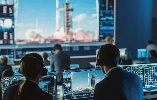 deux personnes sur des moniteurs dans la salle de contrôle de mission regardant un lancement