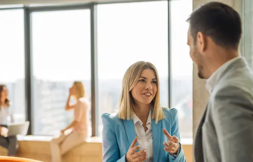 Ung kvinnlig och manlig kollega står och samtalar i en öppen yta på kontoret medan två kollegor syns sittande i bakgrunden