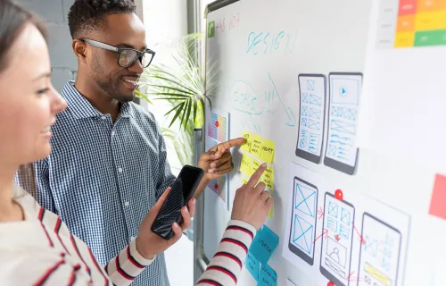 Två personer samarbetar vid en whiteboard med en mobilapp-design. De pekar på skisser och post…