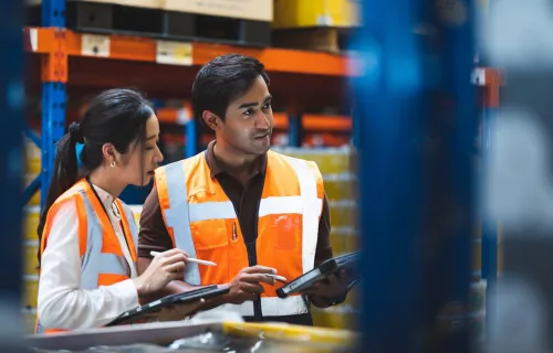 A warehouse worker and manager check stock and inventory using a digital tablet in a retail warehouse full of shelves containing goods.