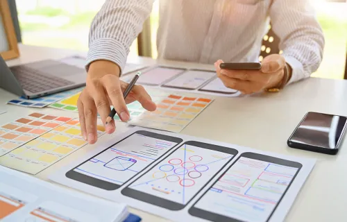 "En person som arbetar med mobilapp-design, med smartphoneskisser och wireframes utspridda på ett skrivbord. Personen håller en mobiltelefon i ena handen och en penna i den andra, medan en bärbar dator och färgprover syns i bakgrunden.