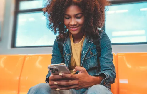 Junge Frau sitzt in einer U-Bahn und schaut lächelnd auf ihr Iphone