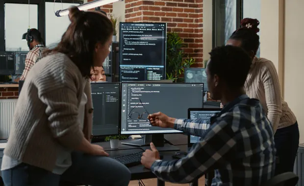 Group of professionals sat at a desk looking at a computer screen which shows coding 