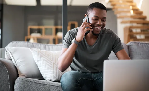 En man sitter i en soffa och pratar i telefon samtidigt som han använder en laptop. Han ler och ser avslappnad ut, omgiven av modern inredning med trappa och hyllor i bakgrunden.