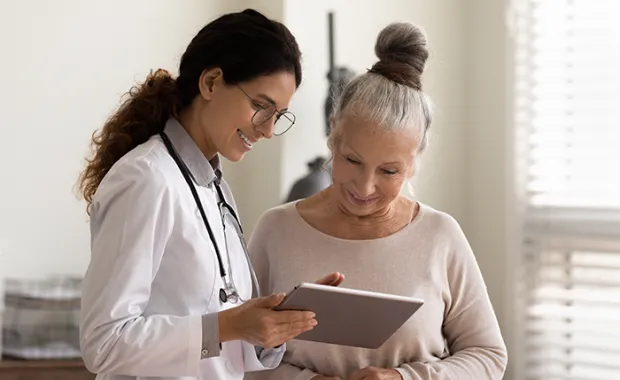 Happy female doctor and senior patient look at tablet screen discuss treatment or therapy on gadget.