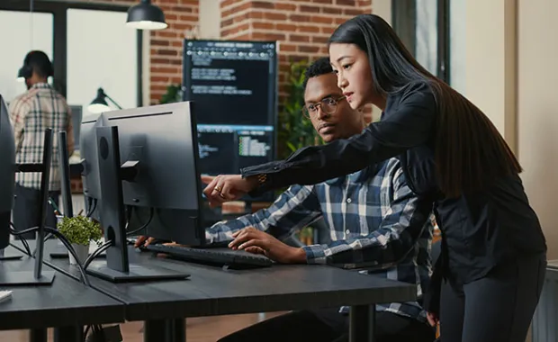 Two programers holding laptop with coding interface walking towards desk and sitting down talking about online cloud computing