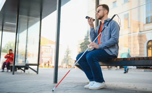 Junger blinder Mann mit Smartphone sitzt auf einer Bank und spricht.