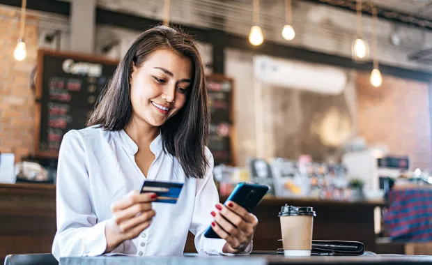 Kvinna sitter på ett cafe med sitt kreditkort i ena handen och smarphone i andra