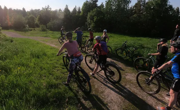 En grupp människor på mountainbikes stannar på en grusväg omgiven av gröna ängar och träd. De verkar ta en paus eller diskutera något, medan solen skiner genom träden i bakgrunden.