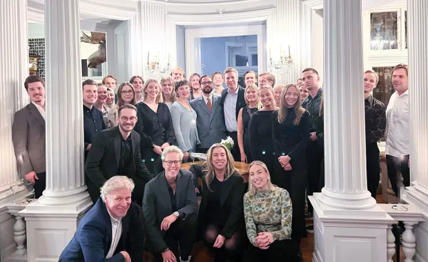 Gruppbild på en större samling personer i formella kläder som står och sitter i ett elegant rum med höga vita pelare och kristallkronor i bakgrunden. Personerna ler mot kameran och ser ut att vara samlade för en formell eller festlig tillställning.