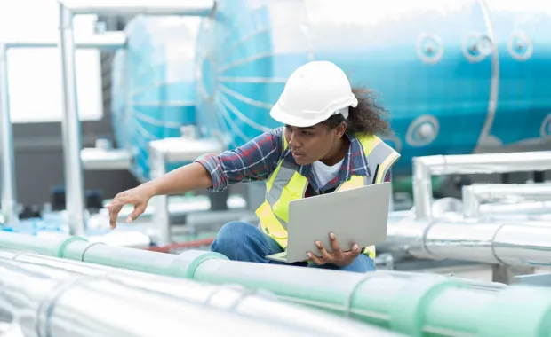 engineer wearing a hard hat inspecting pipes in a plant with a tablet