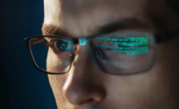 Bedragare som arbetar vid datorn, där man ser koden från skärmen lysa i hand glasögon
