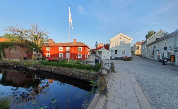 En pittoresk liten stadsmiljö med röda och vita trähus längs en kullerstensgata. En liten å med spegelblankt vatten rinner bredvid gatan, och en flaggstång med en hissad flagga syns i bakgrunden under en klar blå himmel.