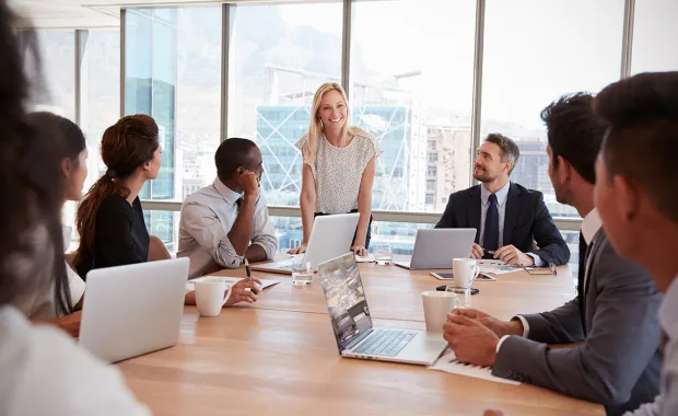 En grupp kollegor som sitter vid ett konferensbord under ett möte. En kvinna står upp och pratar, medan andra lyssnar. Laptops och kaffekoppar syns på bordet, och stora fönster i bakgrunden släpper in naturligt ljus.