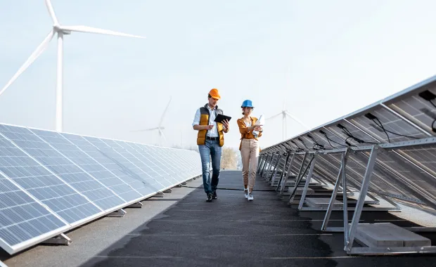 engineers in PPE walking between solar panels with wind turbine in background