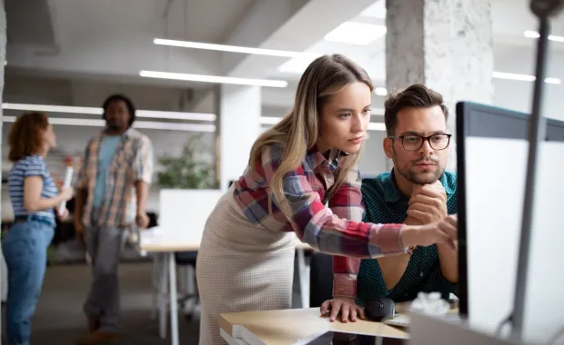 A person pointing at a computer screen while talking to a colleague