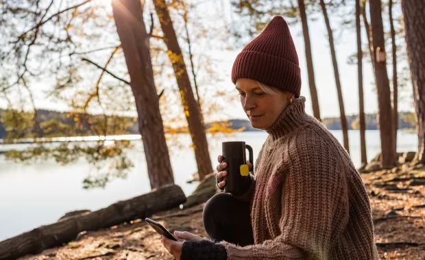 A person sitting lakeside looking at a phone with a mug in hand