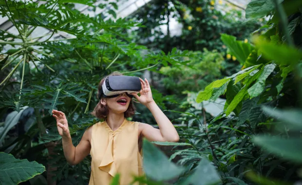 Person wearing VR goggles standing inside a greenhouse and surrounded by plants.