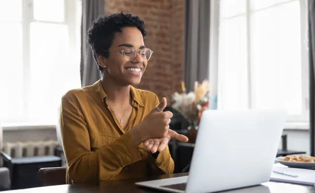 Lächelnde Frau mit Brille sitzt vor einem Laptop und führt ein Videogespräch und nutzt dabei Gebärdensprache für eine barrierefreie Kommunikation