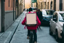 En kille cyklar med en stor ryggsäck i en smal citygränd där andra människor ses gående och bilar står parkerade längs husfasaderna