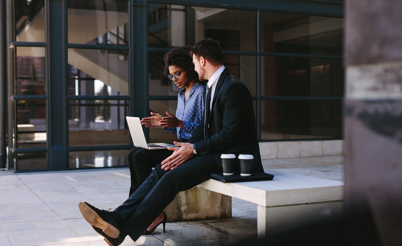 Two people sitting on a bench talking while looking at a computer