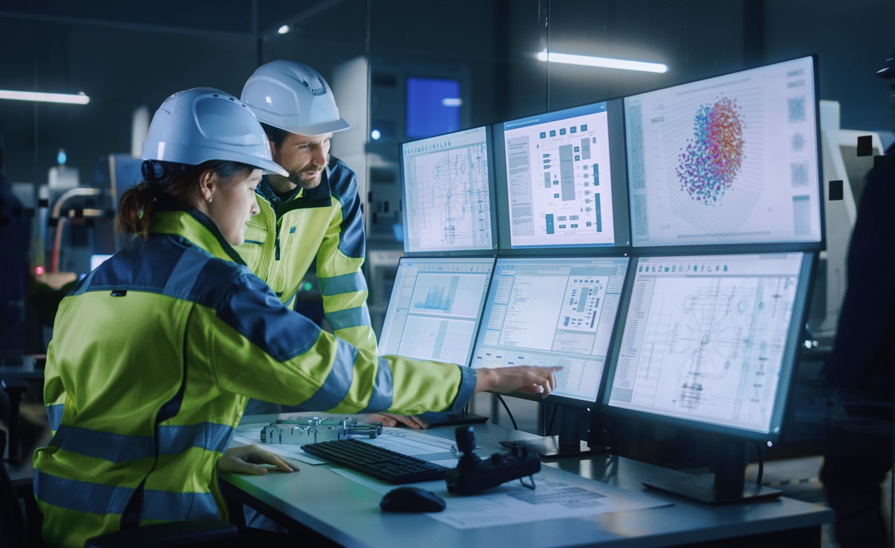 Two people wearing hard hats looking at multiple computer screens