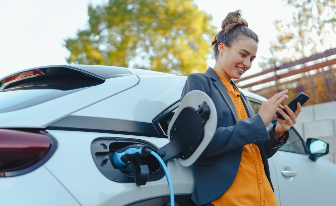 woman with smartphone waiting while electric car charges in home-charging-station